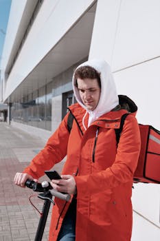 Young delivery man using phone while riding electric scooter in urban setting.